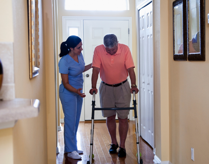 2 Female nurse helping man with walker move through hallway in house.