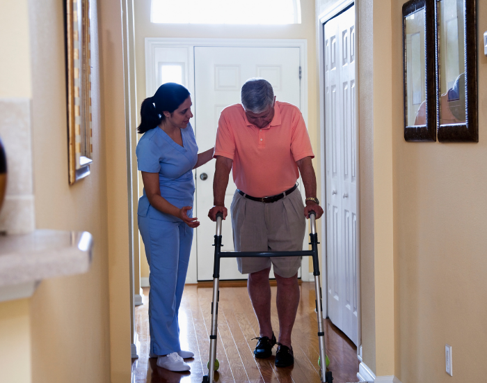 Female nurse helping man with walker move through hallway in house.